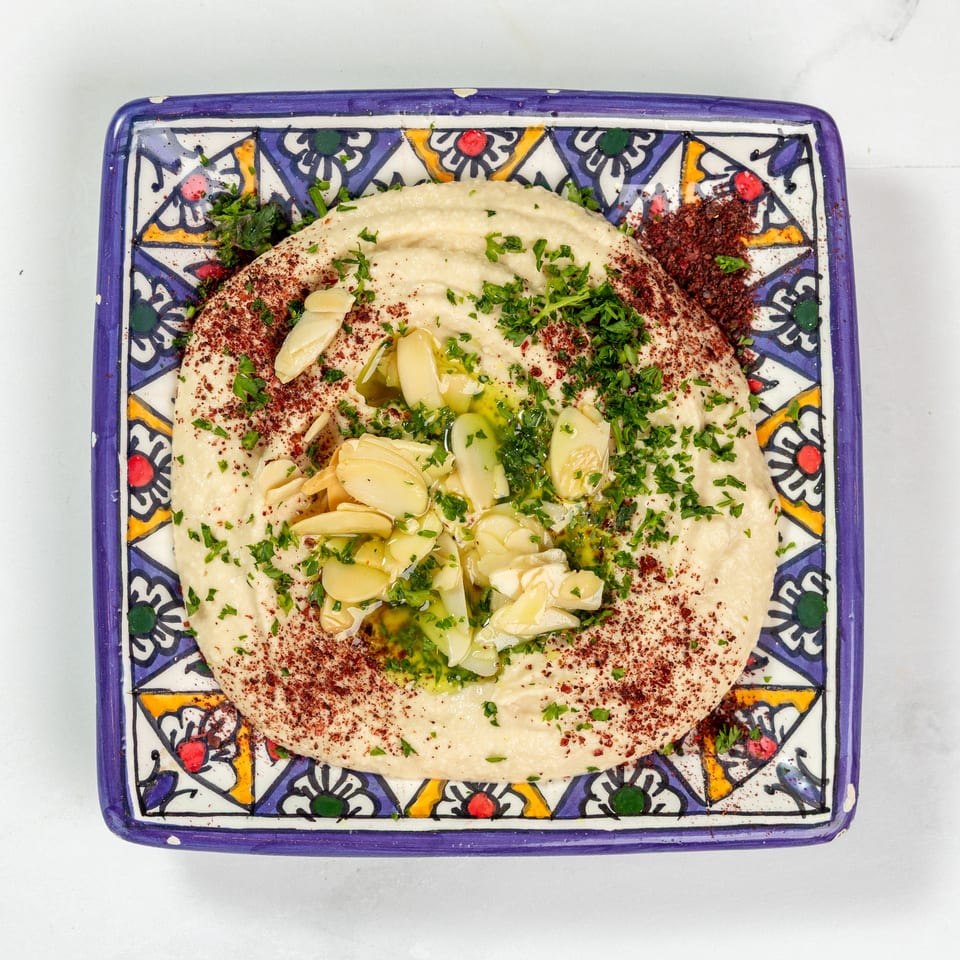 Creamy hummus topped with sliced garlic, parsley, sumac, and olive oil on a decorative plate.
