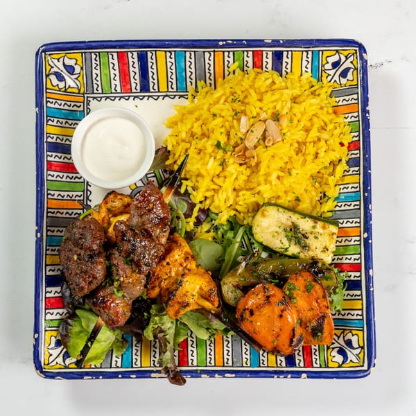 A colorful plate of grilled chicken and beef skewers, yellow rice, roasted vegetables, and a side of white sauce.
