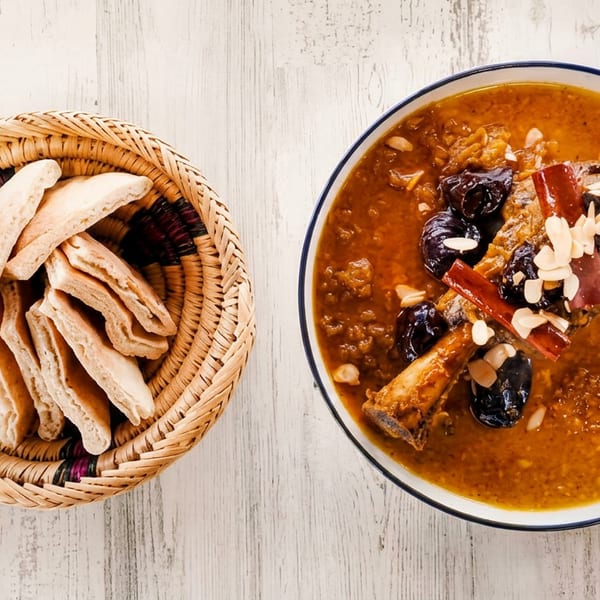 A bowl of Moroccan tagine with lamb, prunes, almonds, and cinnamon sticks, next to a basket of flatbread.