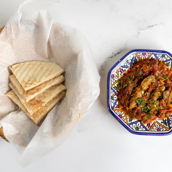 A colorful dish of stewed vegetables with herbs next to a basket of pita bread.