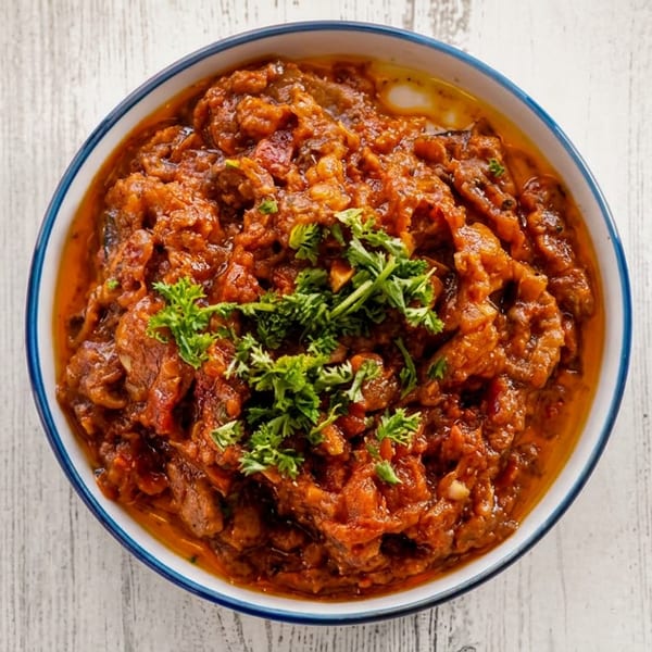 A bowl of rich, red-orange stew, garnished with fresh green parsley, on a white wooden table.