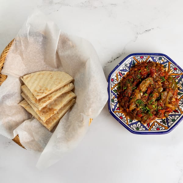 A basket of pita bread and a colorful plate of stewed vegetables with herbs on a white marble surface.