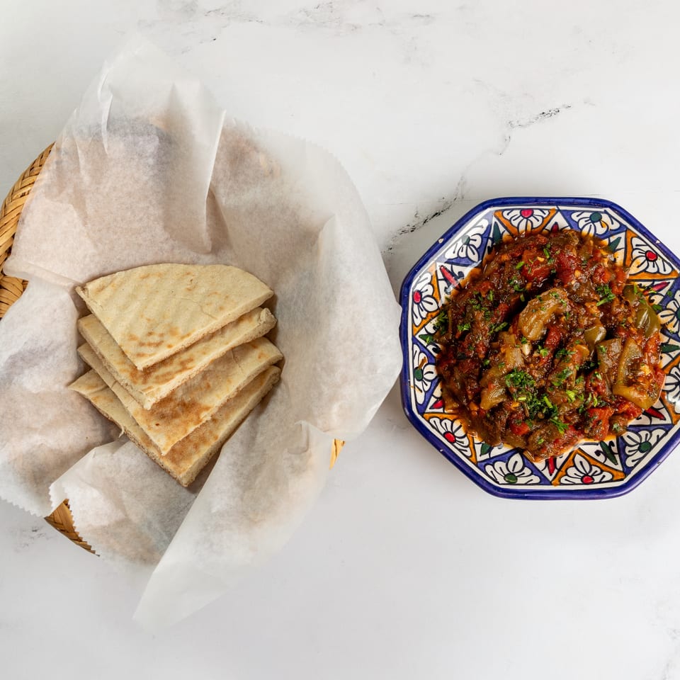 A basket of pita bread and a colorful plate of stewed vegetables with herbs on a white marble surface.