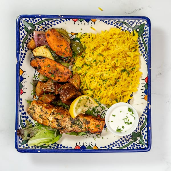 Grilled salmon with roasted vegetables, yellow rice, a lemon wedge, and a side of white sauce.